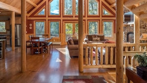 A spacious wooden cabin with a high ceiling, large windows, a dining area, a living room, and a stone fireplace. - Rediker Retreat- Authentic Riverfront Luxury Log Home (Newry)