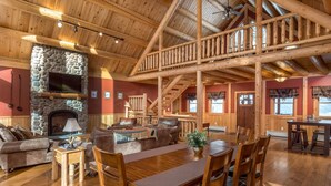 A spacious lodge with a stone fireplace, wooden dining area, and a staircase leading to an upper level. - Rediker Retreat- Authentic Riverfront Luxury Log Home (Newry)