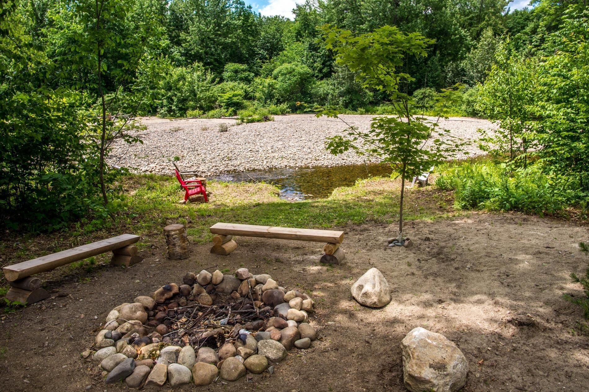 A campsite with a fire pit surrounded by rocks, two wooden benches, and a red chair.