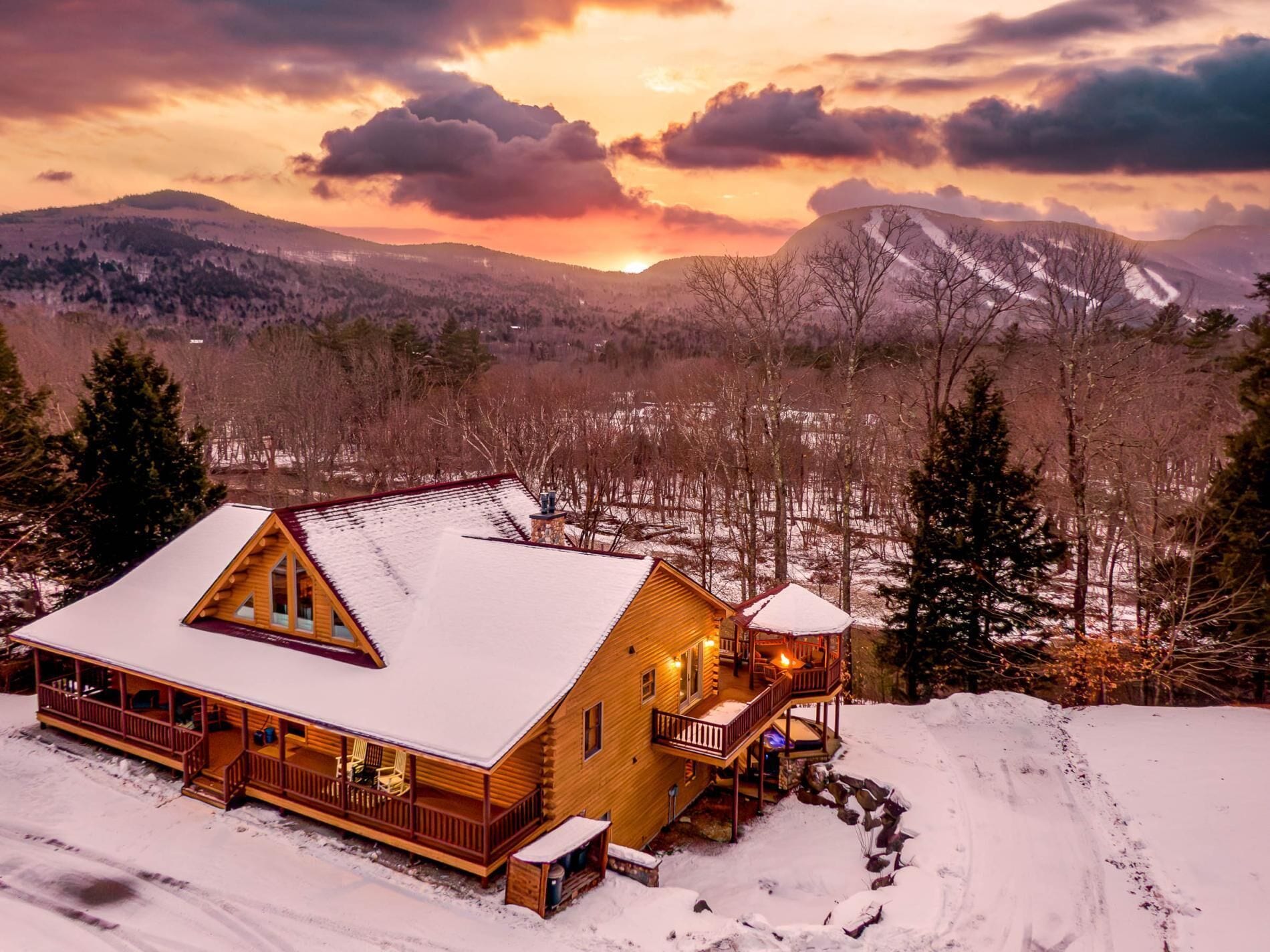 A log cabin with a snow-covered roof, a deck, and a stone fireplace.