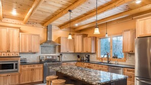 A modern kitchen with wooden cabinets, a large island with stools, stainless steel appliances, and a window with a view of trees. - Rediker Retreat- Authentic Riverfront Luxury Log Home (Newry)
