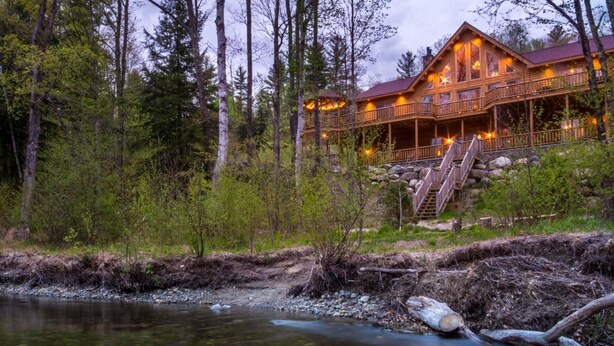 A lodge with a wooden exterior, a stone foundation, and a balcony overlooking a river. - Rediker Retreat- Authentic Riverfront Luxury Log Home (Newry)