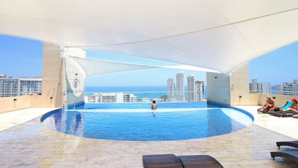 Piscina infinita con vistas al océano y al horizonte de la ciudad, rodeada de sillones de descanso y un área cubierta.