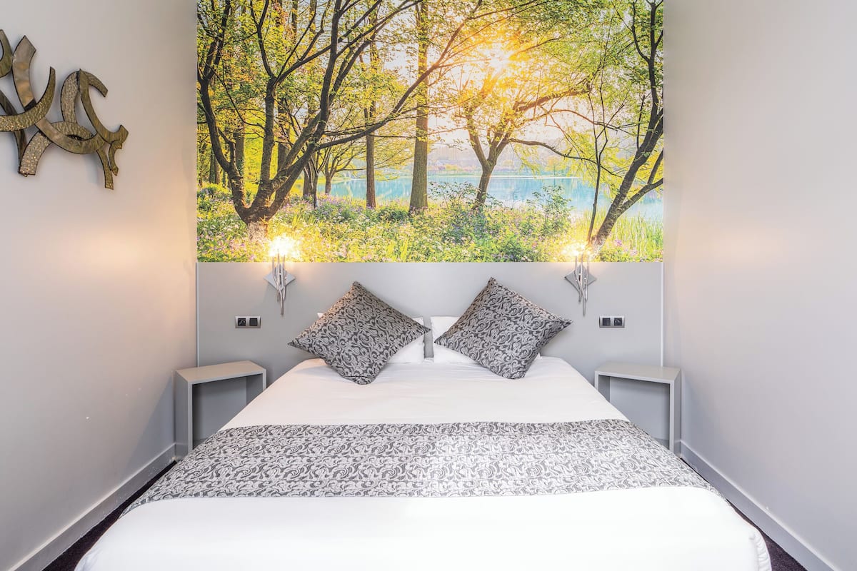 A bedroom with a large bed, two bedside tables, a wall-mounted artwork of a forest scene, and two wall-mounted lights.