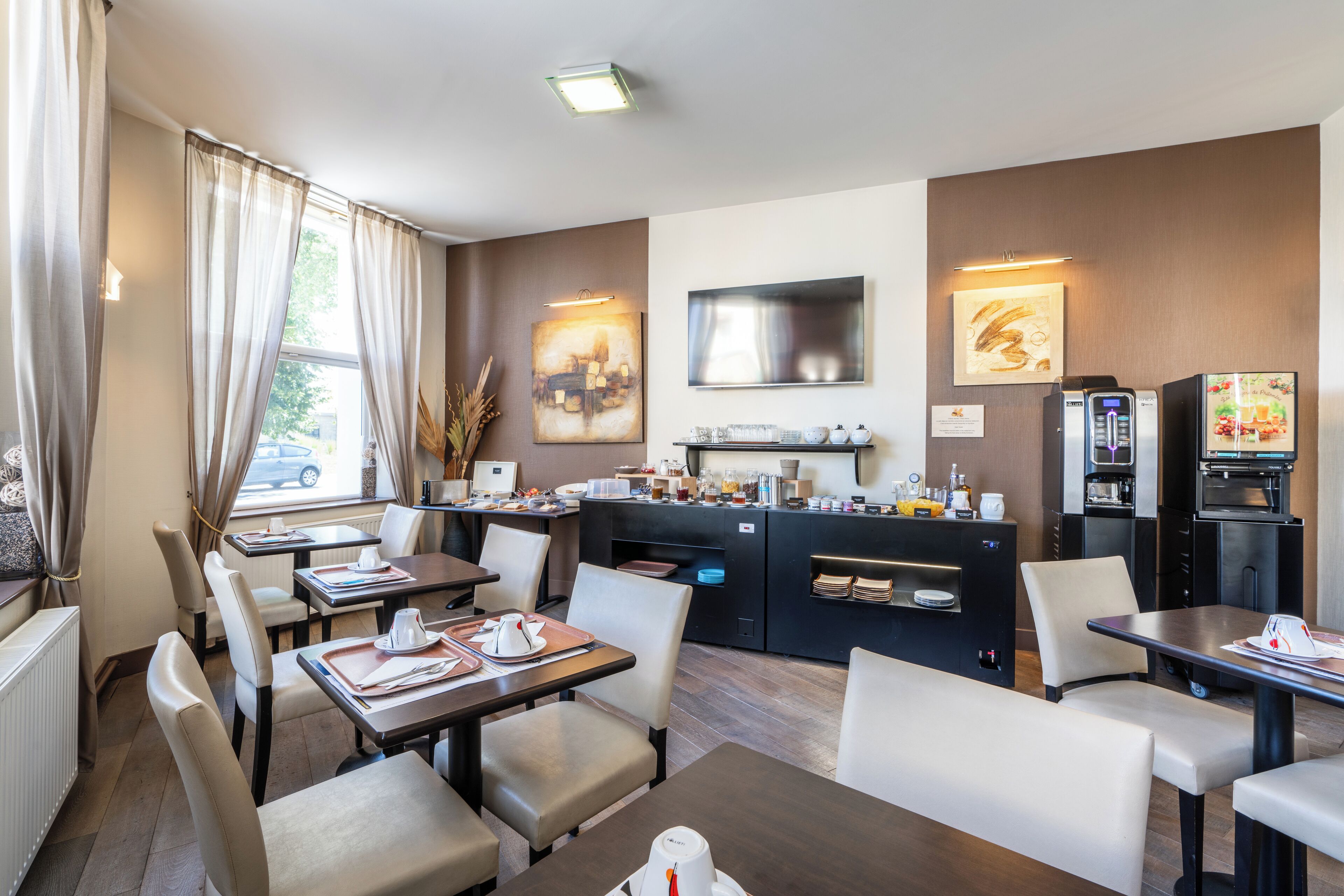 A modern hotel dining area with tables, chairs, a kitchen area, and a TV mounted on the wall.
