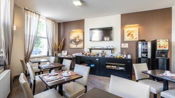 A modern hotel dining area with tables, chairs, a kitchen area, and a TV mounted on the wall.