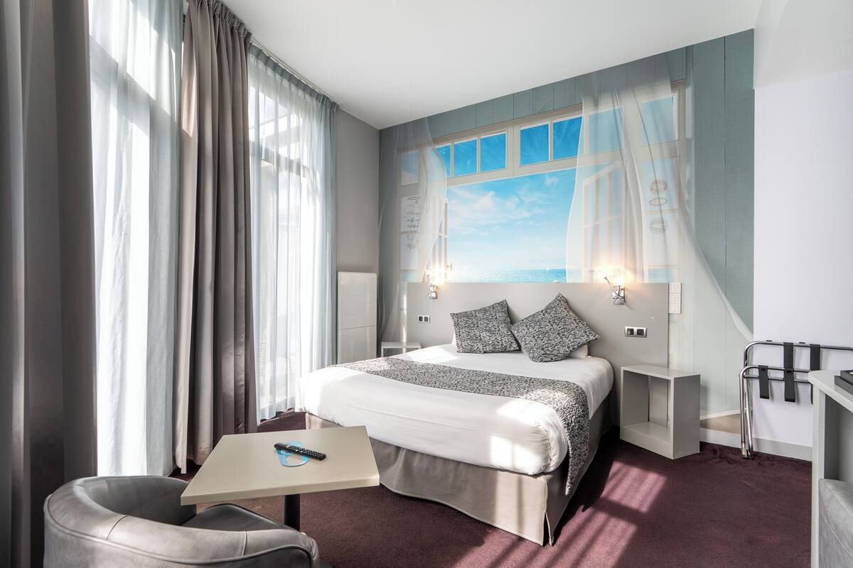 A modern hotel room with a large bed, a desk, and a chair. There is a window with curtains, a nightstand, and a wall-mounted lamp.