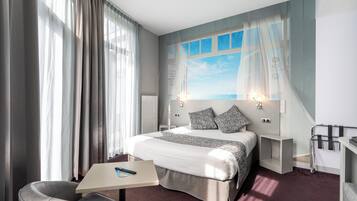 A modern hotel room with a large bed, a desk, and a chair. There is a window with curtains, a nightstand, and a wall-mounted lamp.