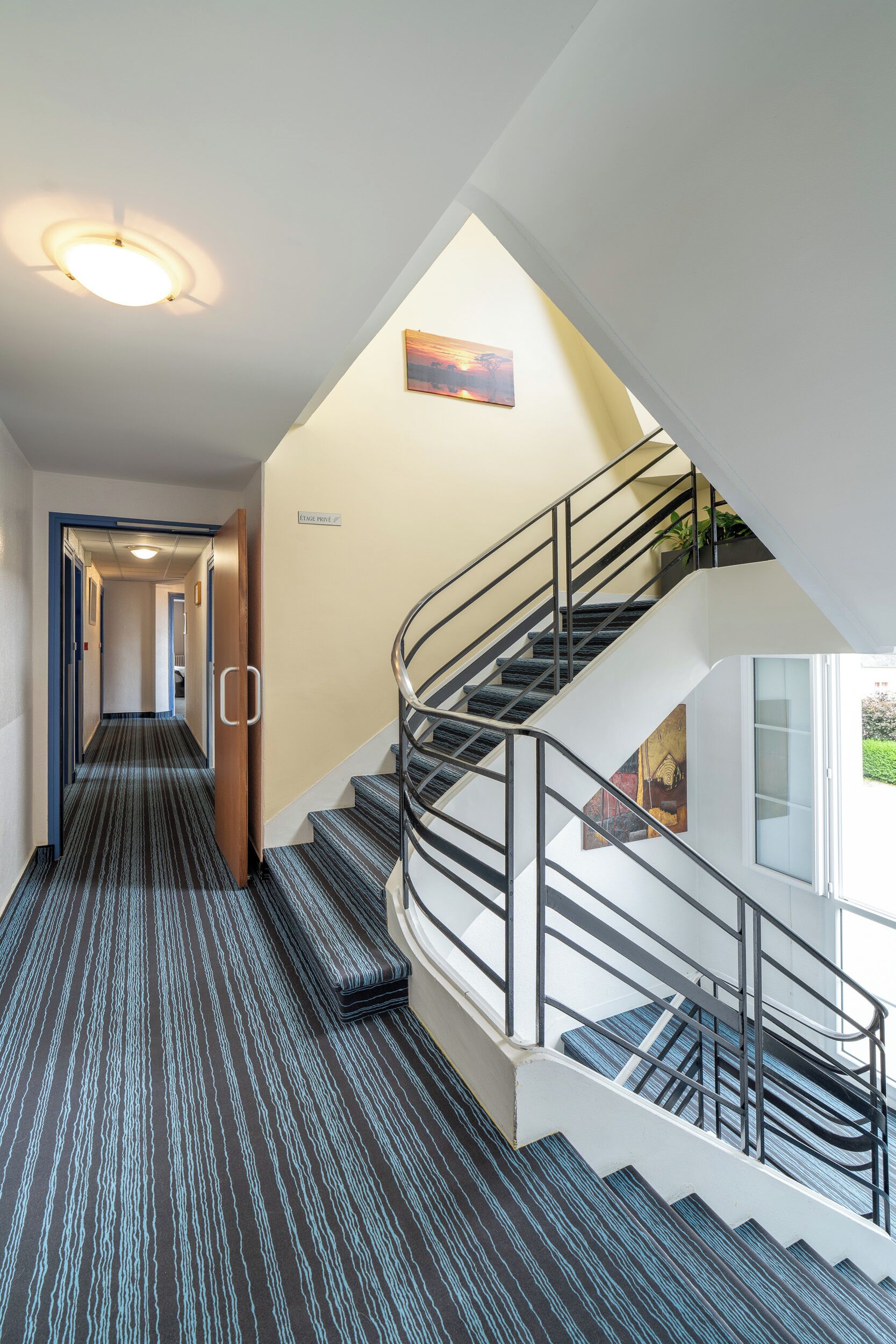 Een modern hotelinterieur met een beklede trap, een gang die naar kamers leidt, en een schilderij aan de muur.