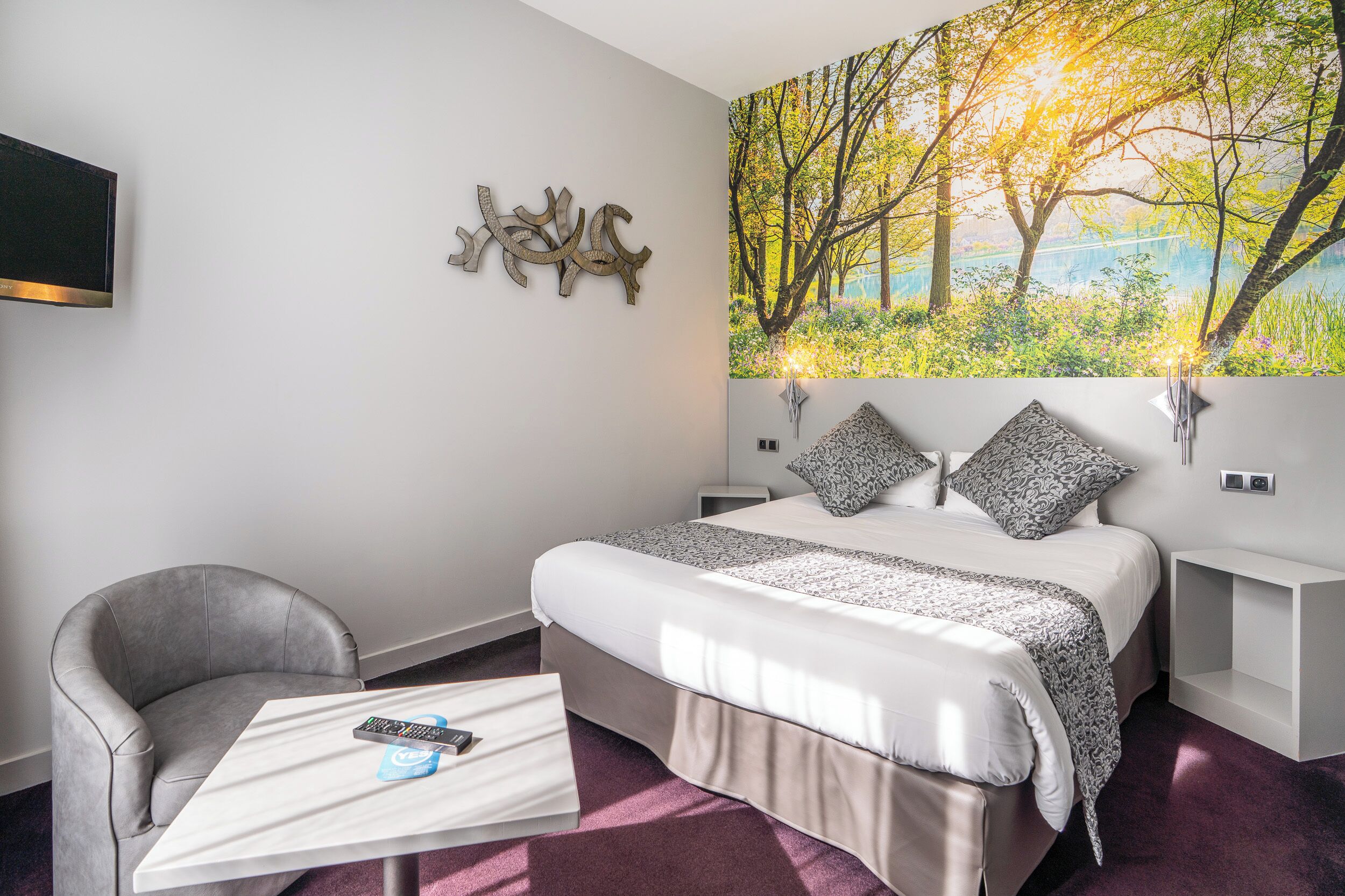 A bedroom with a bed, a chair, a table with a remote, a wall-mounted TV, and a painting of trees.