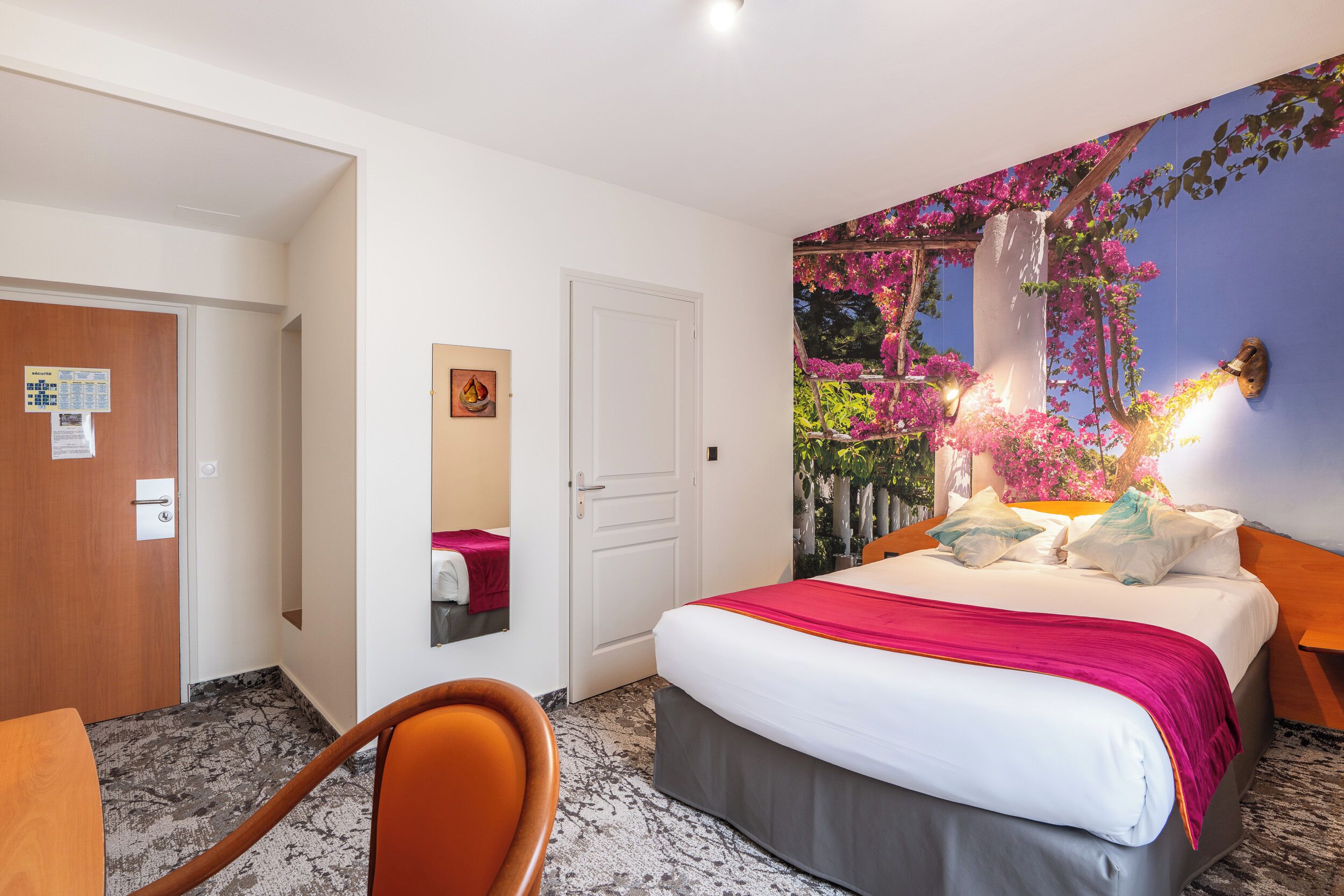 Habitación de hotel con cama, escritorio y silla. Se ve una puerta y una pared con un mural floral.