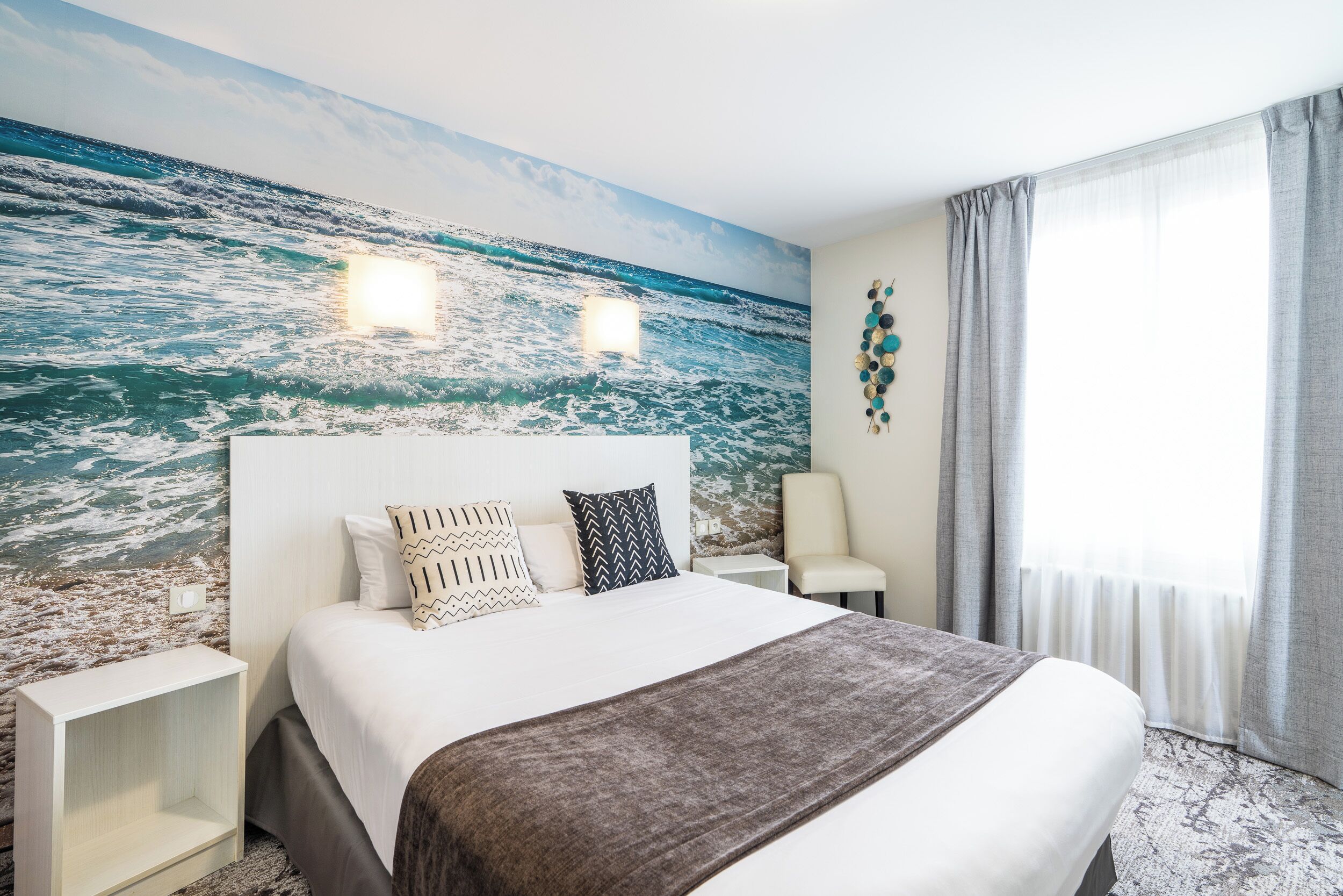 Une chambre d’hôtel avec un lit, une table de chevet, une chaise et une grande fresque représentant l’océan sur le mur.