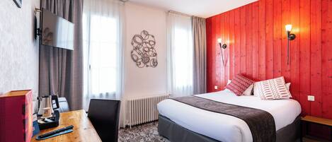 A hotel room with a bed, a desk with a remote control, a wall-mounted TV, and a red accent wall.