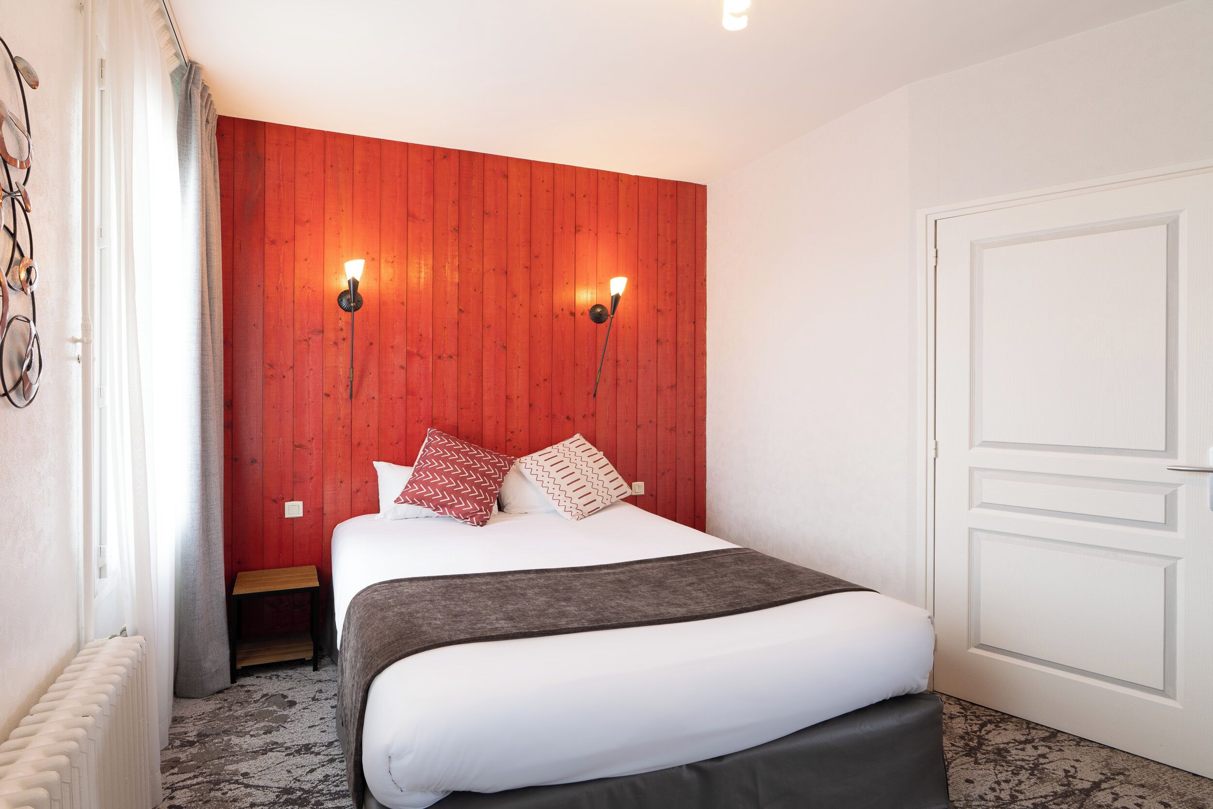 Une chambre d’hôtel avec un mur d’accent en bois, un lit avec une literie blanche et une couverture grise, deux lampes fixées au mur et une porte blanche.