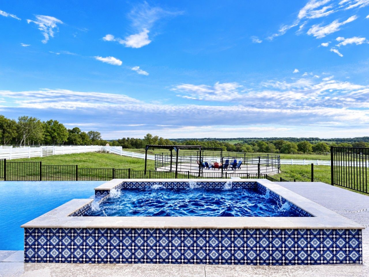 A rectangular hot tub with a patterned tile border, surrounded by a fence, with a view of a grassy area and trees in the background.