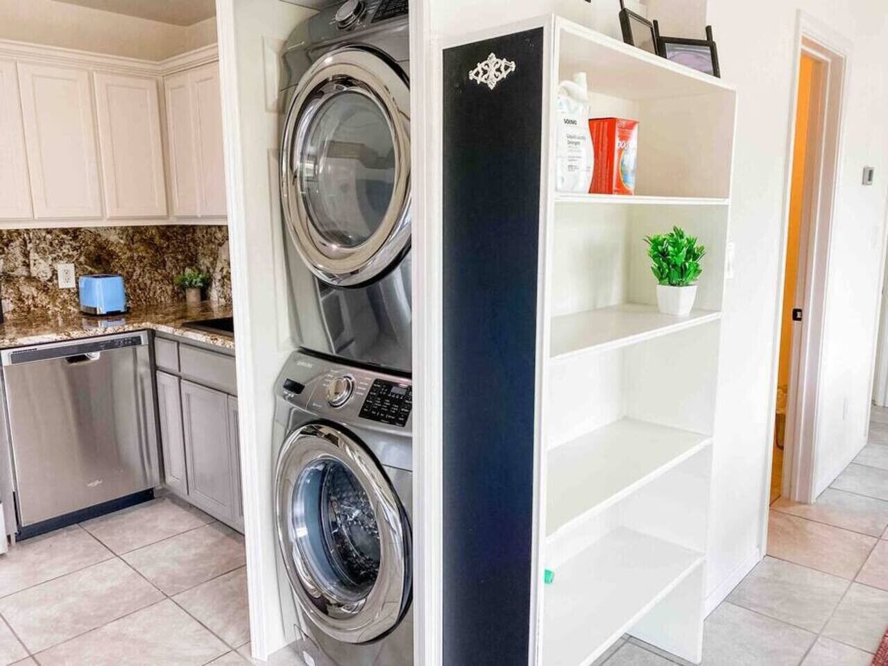 Une cuisine équipée d’un lave-linge et d’un sèche-linge intégrés, d’un lave-vaisselle et d’une étagère blanche où est posée une plante en pot.
