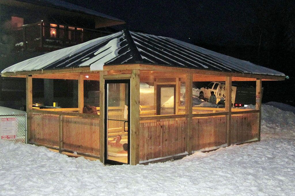 Une cabane en bois avec un toit en métal, entourée de neige, dotée d’une porte et de fenêtres en verre.