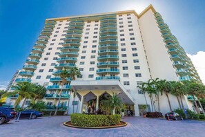 A high-rise hotel with multiple balconies, a central entrance, and palm trees in the foreground.