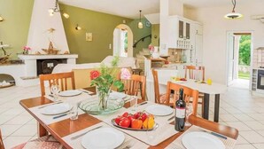Uma sala de jantar com uma mesa de madeira posta para quatro pessoas, rodeada por cadeiras. Há um vaso com flores sobre a mesa, além de uma garrafa de vinho e um copo. O quarto tem uma parede verde e uma lareira com objetos decorativos.