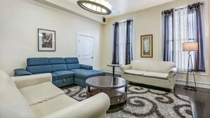 Un salón con un sofá modular, una mesa de centro redonda y un sillón color beige. Se observa una puerta y una ventana con cortinas.