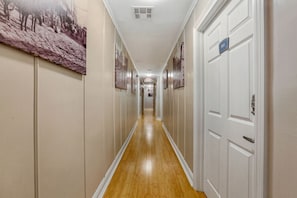 Un long couloir étroit, doté d’un plancher en bois et de murs blancs. Plusieurs portes se trouvent sur le côté droit, chacune étant surmontée d’un numéro.