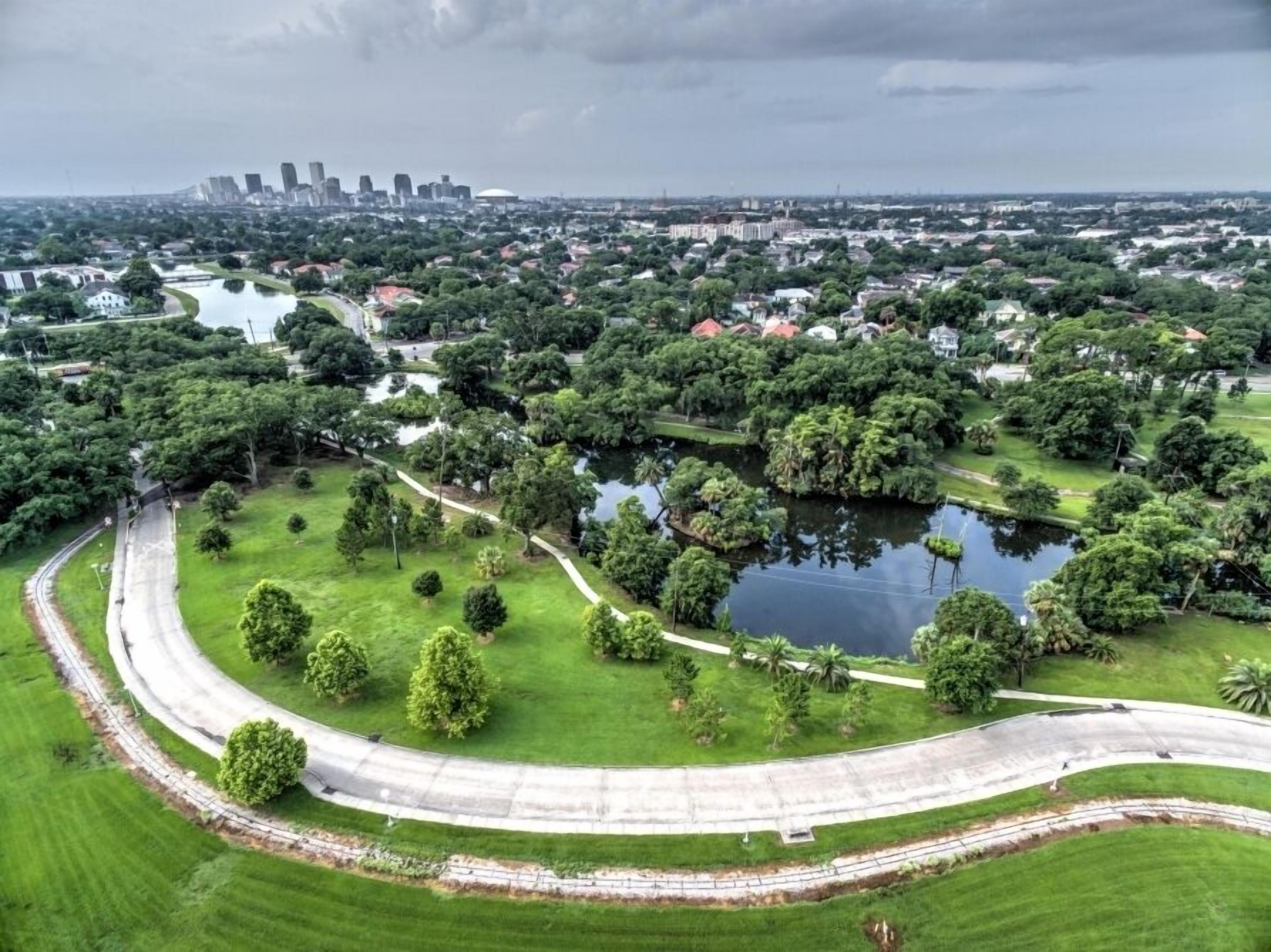 An aerial view of a park with a curved road, a lake, and a city skyline in the background.