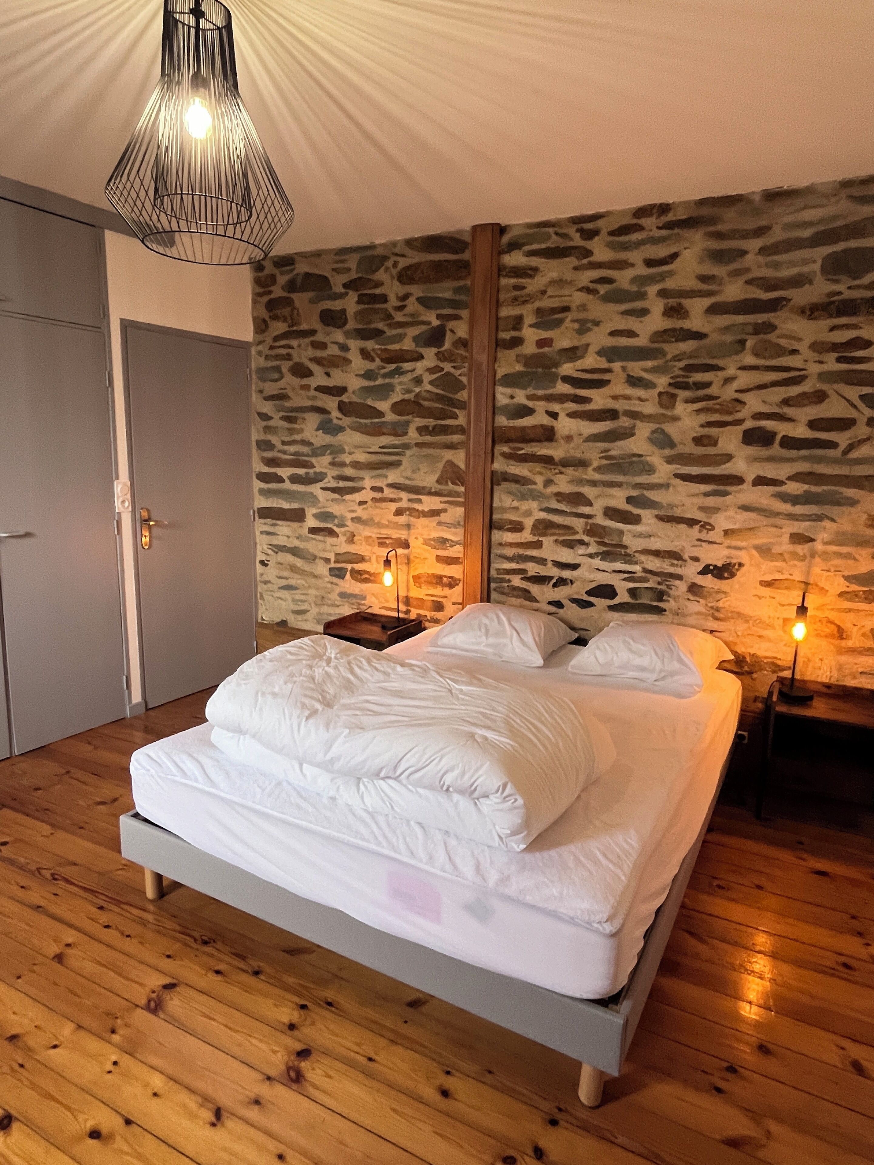 Une chambre avec un mur en pierre, un plancher en bois, un lit avec une literie blanche et deux lampes de chevet.