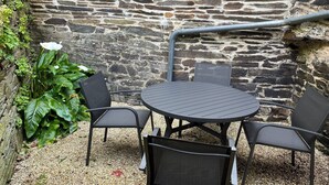 Un muro de piedra con una tubería metálica, una mesa redonda con cuatro sillas y plantas verdes.