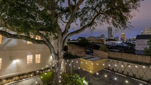 Un patio arbolado con un edificio blanco, luces colgantes y un horizonte urbano al fondo.