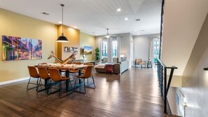 A spacious living area with a dining table, chairs, and a sofa. There are paintings on the walls and a staircase leading to another level.