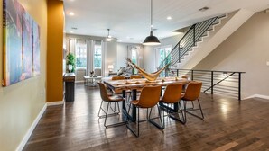 A spacious dining area with a large wooden table and modern chairs, a staircase with a black railing, and a view of the outdoors through large windows.