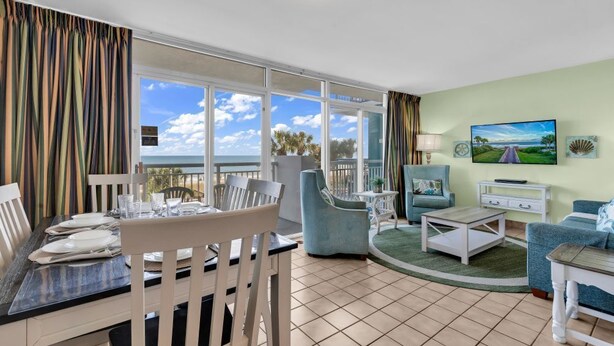 A room with a dining table set for four, a sofa, a TV, and a balcony with a view. - Floor to Ceiling Windows at Atlantica Resort 🍹Oceanfront 🍹 (Myrtle Beach)