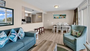 A living room with a sofa, a coffee table, a dining table with chairs, a kitchen area, and a large window with a view. - Floor to Ceiling Windows at Atlantica Resort 🍹Oceanfront 🍹 (Myrtle Beach)