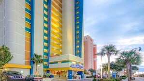 A multi-story hotel with a prominent yellow and blue facade, a central entrance, and a parking area with cars. - Floor to Ceiling Windows at Atlantica Resort 🍹Oceanfront 🍹 (Myrtle Beach)