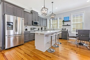 Une cuisine moderne avec des meubles gris, des appareils en acier inoxydable, un îlot central blanc doté de tabourets, et une grande fenêtre avec des stores.