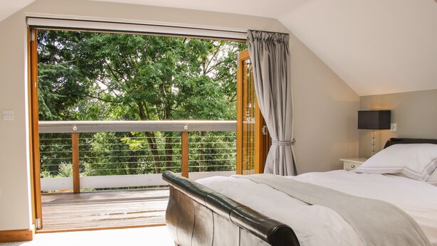 A bedroom with a large bed, a nightstand, a lamp, and a view of a balcony with a railing. - Sunnyside (Leominster)