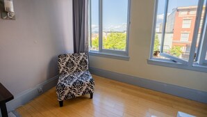 Une pièce dotée d’une grande fenêtre, d’un fauteuil à motifs, d’un plancher en bois et d’un luminaire fixé au mur.