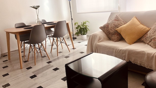 Uma sala de estar com sofá, mesa de centro, mesa de jantar com cadeiras, planta em vaso e abajur.