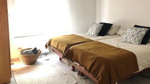 Quarto com duas camas, piso de madeira, cesta de palha e janela com luz natural.