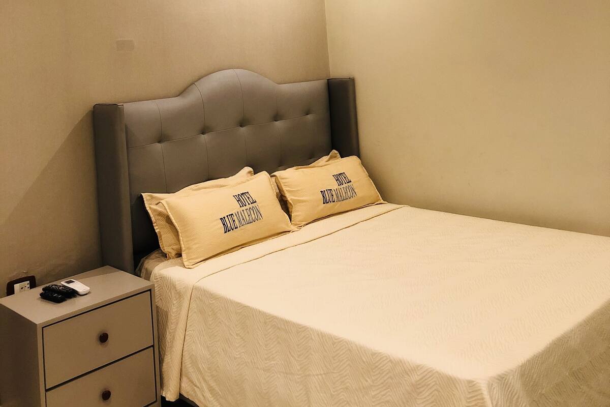 Une chambre à coucher avec un lit, deux oreillers, une table de chevet avec des tiroirs et une lampe fixée au mur.
