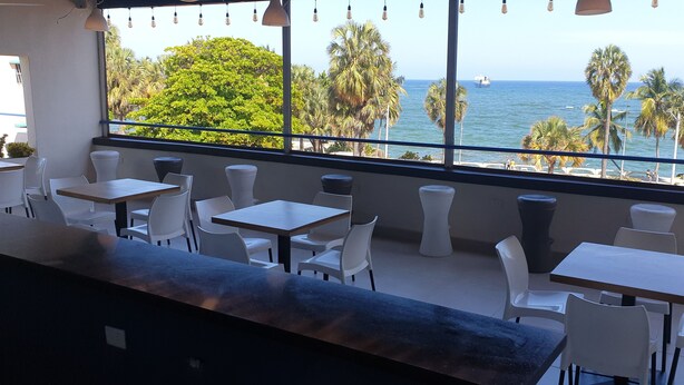 Un restaurant doté d’une terrasse offrant une vue sur la mer, avec des chaises et des tables blanches, et une vue sur les palmiers et les bateaux.