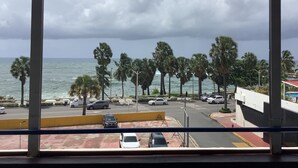 Vue depuis l’intérieur d’un bâtiment, donnant sur un parking où l’on aperçoit des voitures, des palmiers et l’océan, le tout sous un ciel nuageux.