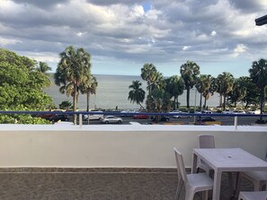 A balcony with a view of the sea, palm trees, and parked cars. - Hotel Blue Malecón (Santo Domingo)