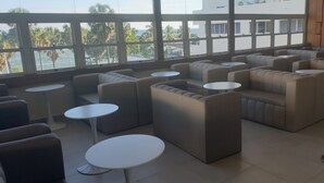 Un espace de détente moderne, avec des canapés et des tables rondes, de grandes fenêtres offrant une vue sur les palmiers et les immeubles, et un mur en bois agrémenté d’une touche de vert.