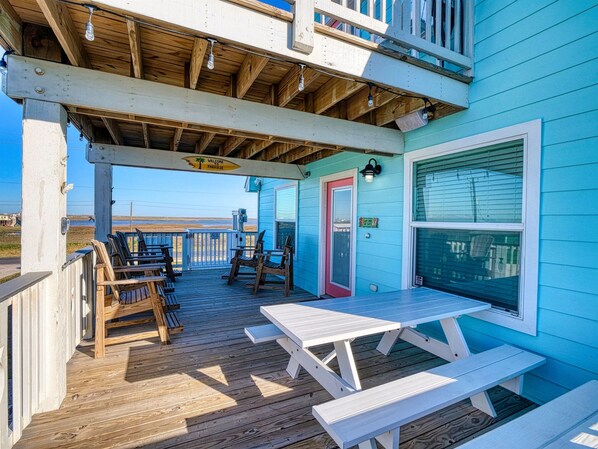 Una terraza de madera con mesas y sillas de picnic, un espacio cubierto con un letrero que dice 'Welcome Paradise', y vista a la playa.