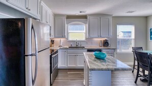 A modern kitchen with white cabinetry, a stainless steel refrigerator, a granite countertop, and a dining area with a wooden table and chairs. - Modern A-Frame | 3BR/2BA | Pet-Friendly | Steps to Beach | Huge Deck (Freeport)