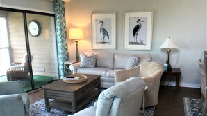 Ein Wohnzimmer mit Sofa, Sesseln, einem Couchtisch und einem gemusterten Teppich. Zwei Wandleuchten und Bilder von Vögeln in Rahmen sind ebenfalls zu sehen.