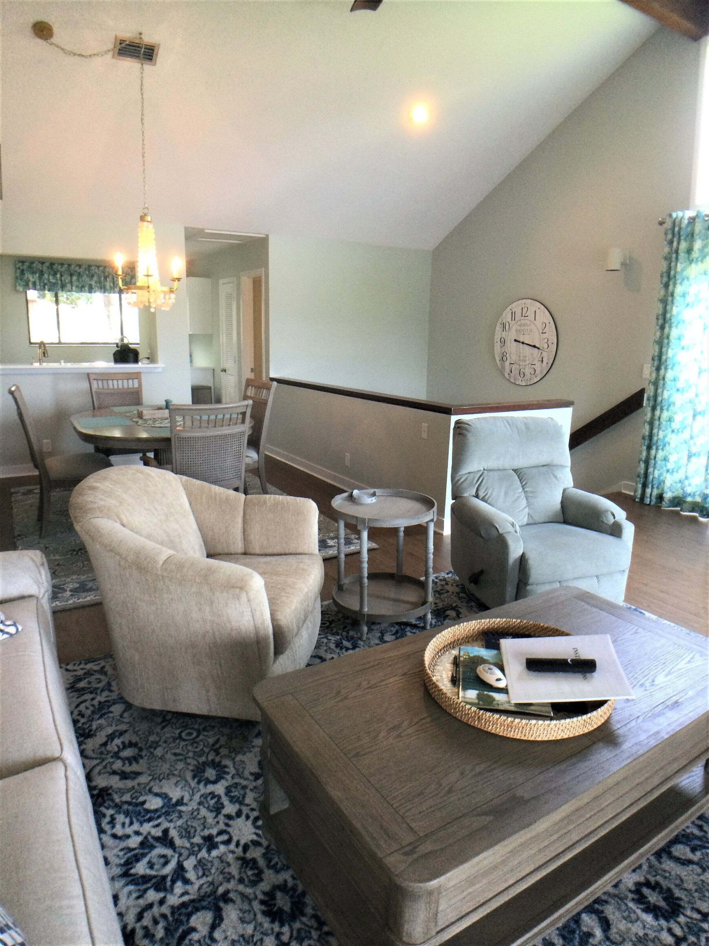 A living room with a sofa, armchair, and a coffee table. There is a dining area with a table and chairs, and a kitchen in the background.