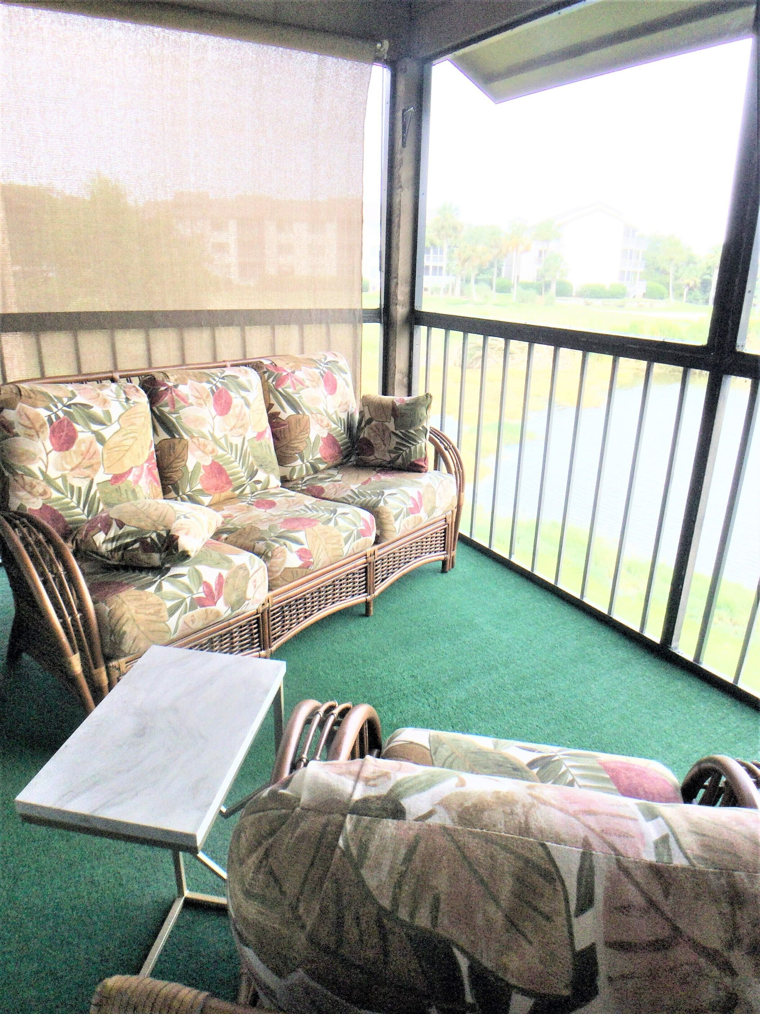 A covered outdoor seating area with a wicker sofa and a small table, surrounded by a green carpet.
