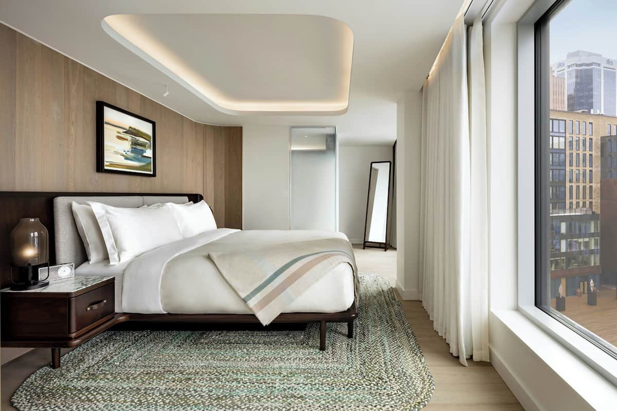 A modern hotel room with a large bed, a nightstand, a mirror, and a view of the city through large windows.
